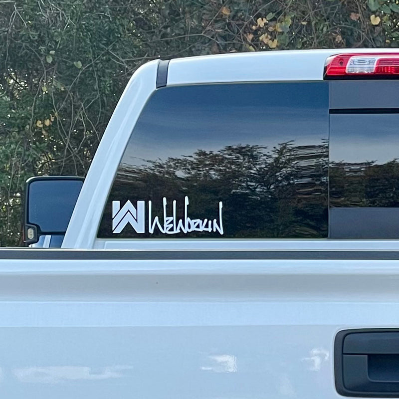 Cropped in image of a WeWorkin Icon/script Logo Direct Transfer window decal in white, on rear window (lower left) of a Chevy truck. Foliage in background.