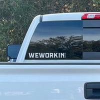 Cropped in image of a WeWorkin Brand text/image Direct Transfer window decal in white, on rear window (lower left) of a Chevy truck. Foliage in background.