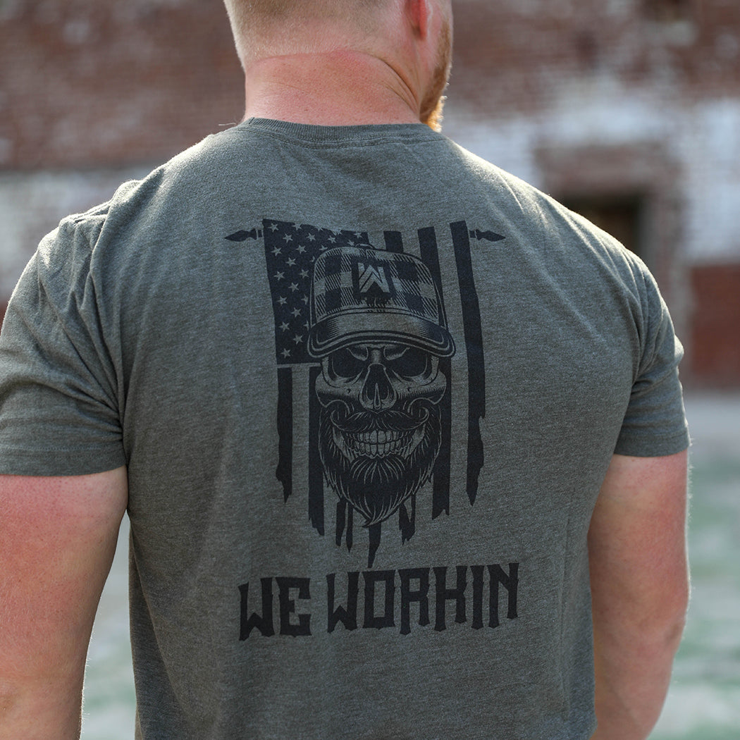 Man pictured from back wearing a We Workin graphic tee in Heather Military Green color. Short sleeve shirt imprinted with a WeWorkin designed graphic—the text "WE WORKIN" in bold, below a graphic bearded skull layered on top of a tattered US flag hanging vertically from a rod (printed in black ink.)