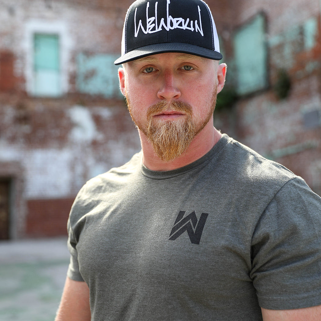 Man pictured from the front wearing a military green WeWorkin tee with the WW icon small on the left chest, imprinted in black ink. He is also wearing the WeWorkin Black/White Retro Trucker hat with WW script logo embroidered large on the front in white thread. 