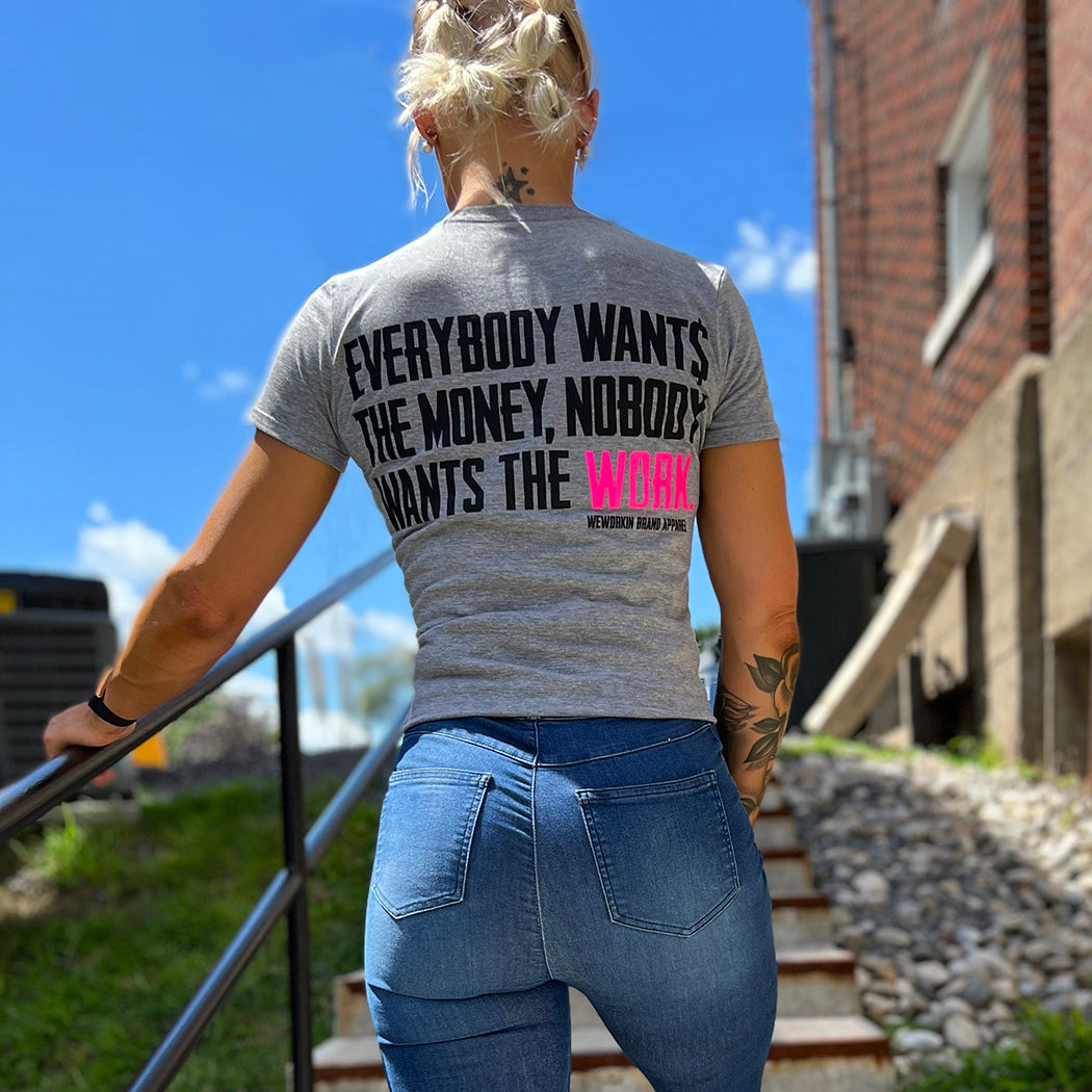 Woman outdoors, pictured from back, wearing jeans and a WE WORKIN Women’s short sleeve tee in aluminum grey. Tee has the popular WEWORKIN tagline "Everybody Wants the Money, Nobody Wants the WORK." printed on the back in black ink, except the word "WORK." is highlighted in flourescent magenta/pink. 