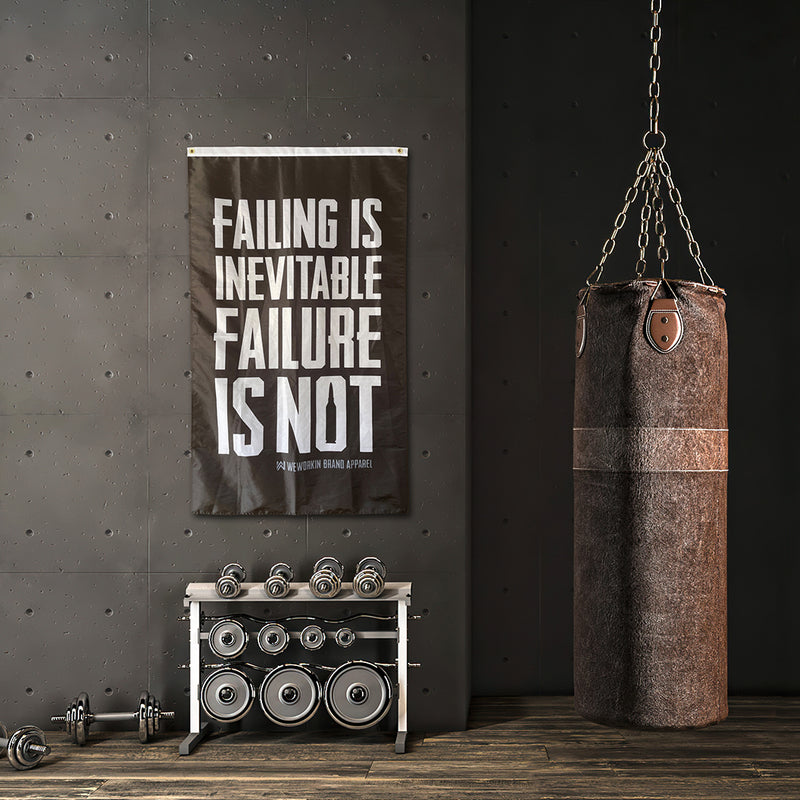WeWorkin Brand "FAILING IS INEVITABLE—FAILURE IS NOT" flag displayed in a commercial gym. Each flag measures approximately 3'W x 5'H. The flag has a black background with all white letters. White, double-stitched, thicker top edge for durability, (2) gold grommets (one at top left and one at top right corners).